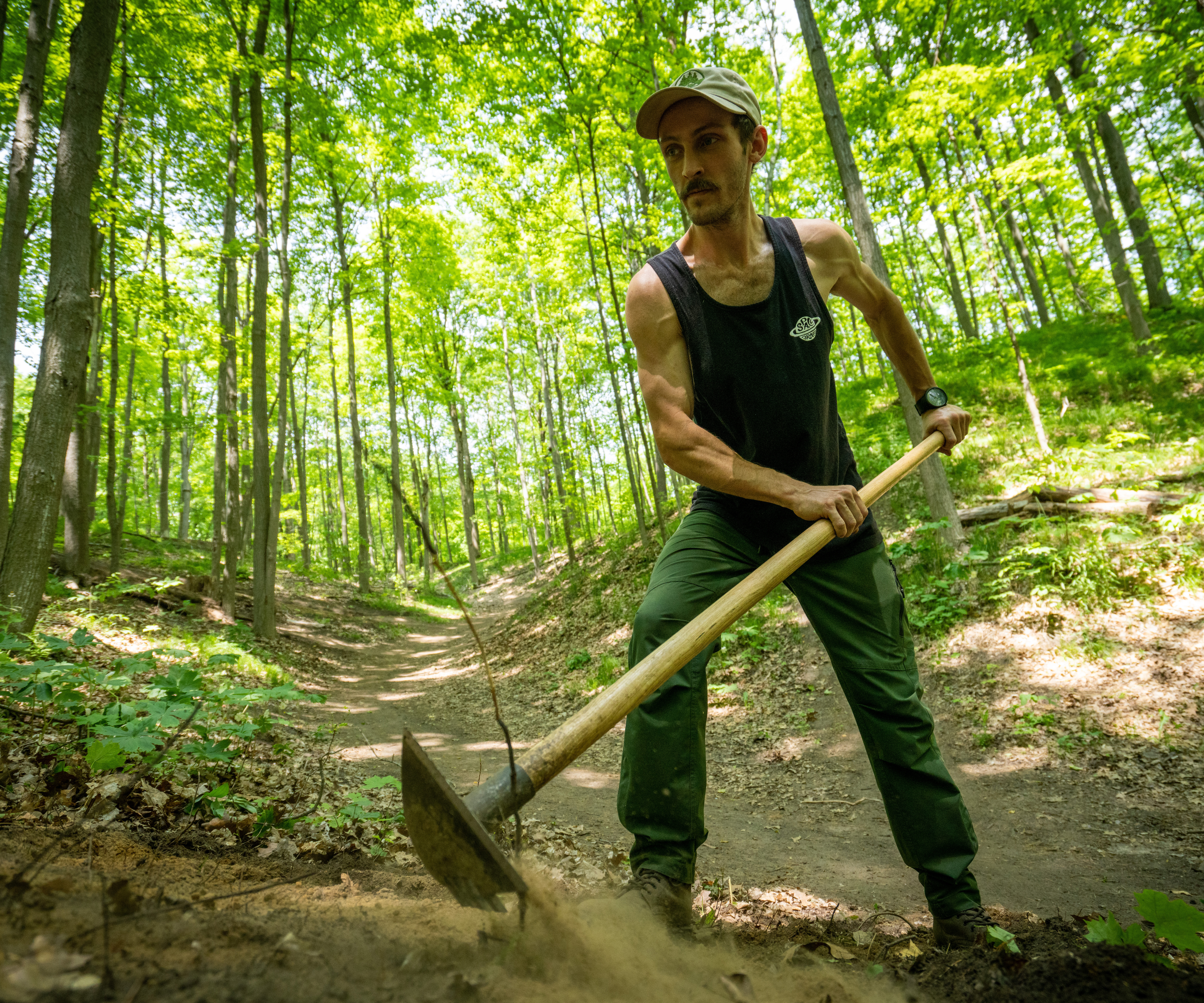 Man raking a trail with green trees in the background.